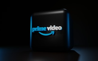 A dark background contrasts with a three-dimensional, illuminated logo in the center. The logo is composed of the words 'prime video' in bold, blue lettering with a distinctive smile-shaped arrow underneath, casting subtle shadows.