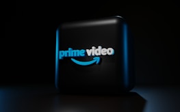 A dark background contrasts with a three-dimensional, illuminated logo in the center. The logo is composed of the words 'prime video' in bold, blue lettering with a distinctive smile-shaped arrow underneath, casting subtle shadows.