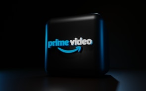 A dark background contrasts with a three-dimensional, illuminated logo in the center. The logo is composed of the words 'prime video' in bold, blue lettering with a distinctive smile-shaped arrow underneath, casting subtle shadows.