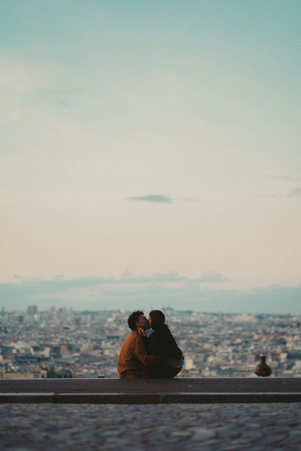 a man and woman kissing on a ledge overlooking a city
