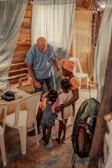 An elderly man in a blue shirt interacts warmly with a group of young children and a woman inside a rustic room. The setting includes curtains and white plastic chairs, creating a cozy and intimate atmosphere.