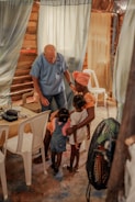 An elderly man in a blue shirt interacts warmly with a group of young children and a woman inside a rustic room. The setting includes curtains and white plastic chairs, creating a cozy and intimate atmosphere.