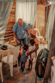 An elderly man in a blue shirt interacts warmly with a group of young children and a woman inside a rustic room. The setting includes curtains and white plastic chairs, creating a cozy and intimate atmosphere.