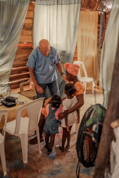 An elderly man in a blue shirt interacts warmly with a group of young children and a woman inside a rustic room. The setting includes curtains and white plastic chairs, creating a cozy and intimate atmosphere.