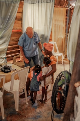 An elderly man in a blue shirt interacts warmly with a group of young children and a woman inside a rustic room. The setting includes curtains and white plastic chairs, creating a cozy and intimate atmosphere.