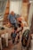 An elderly man in a blue shirt interacts warmly with a group of young children and a woman inside a rustic room. The setting includes curtains and white plastic chairs, creating a cozy and intimate atmosphere.