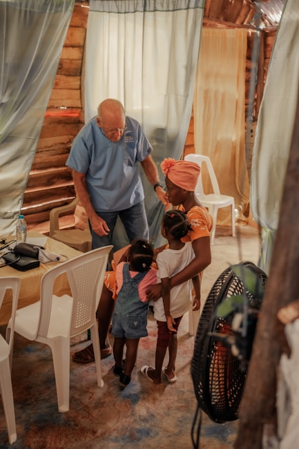 An elderly man in a blue shirt interacts warmly with a group of young children and a woman inside a rustic room. The setting includes curtains and white plastic chairs, creating a cozy and intimate atmosphere.