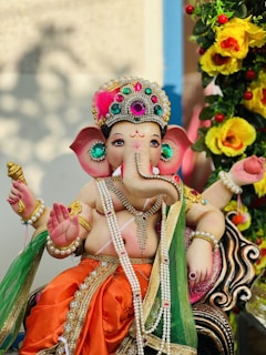 A colorful and intricately designed statue of the Hindu deity Ganesha, adorned with vibrant jewels and dressed in traditional attire. The statue is surrounded by decorative flowers and leaves, enhancing the festive and sacred atmosphere.