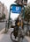 A street scene in an urban area showing a person standing next to a bicycle near an entrance to the Oranienburger U-Bahn station. The person is partially obscured by a pole covered with numerous stickers. The street is flanked by trees and buildings, and there's a tram rail running along the road.