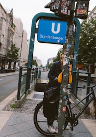 A street scene in an urban area showing a person standing next to a bicycle near an entrance to the Oranienburger U-Bahn station. The person is partially obscured by a pole covered with numerous stickers. The street is flanked by trees and buildings, and there's a tram rail running along the road.