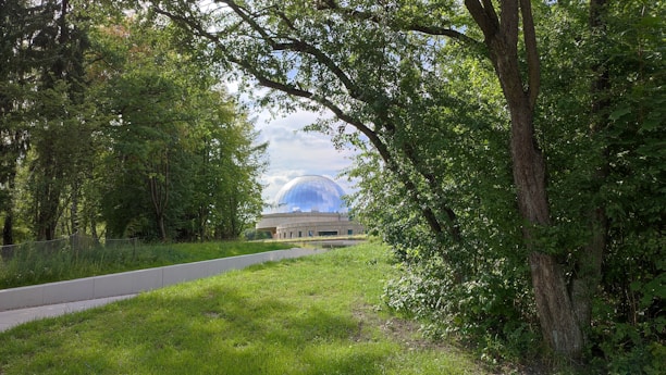 A pathway leads through a lush, green forest clearing toward a building with a large domed structure that reflects a blue sky with some clouds.