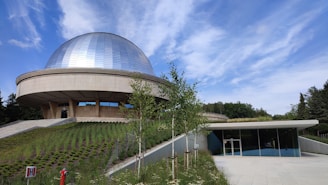 A modern building with a large, shiny dome structure set on a grassy hill. The foreground features a manicured garden with young trees and a paved walkway leading to the entrance of the building. The sky is clear with a few wispy clouds.