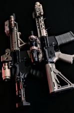 Two tactical rifles are displayed side by side against a dark background. They are equipped with various attachments including scopes and flashlights, featuring a combination of black and tan colors.
