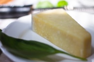 A piece of hard cheese rests on a white plate with a green leaf beside it. The cheese has a wedge shape and a slightly rough texture, indicative of an aged variety. The lighting is soft, creating subtle highlights on the cheese surface, while the background is blurred to focus attention on the cheese and leaf.