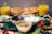 A breakfast spread featuring various food items. In the foreground, there are bowls filled with peanut butter, chocolate spread, and whipped cream, surrounded by fresh blueberries, sliced strawberries, mint leaves, and chocolate chips. In the background, there are two cups of orange juice and waffles on a wooden plate.