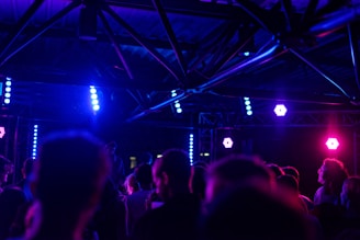 A dark indoor concert or club scene with a group of people facing a stage. Bright purple and blue lights illuminate the space, creating a vibrant atmosphere. Metal trusses and light fixtures are visible above the crowd.