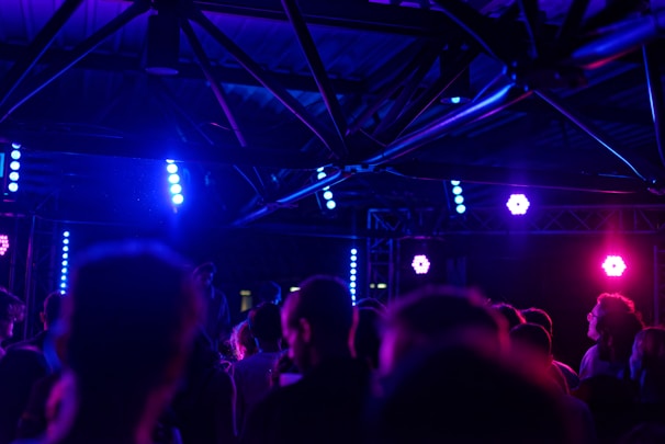 A dark indoor concert or club scene with a group of people facing a stage. Bright purple and blue lights illuminate the space, creating a vibrant atmosphere. Metal trusses and light fixtures are visible above the crowd.