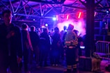 A group of people congregates under an industrial-style roof with metal beams, illuminated by vibrant purple and blue lighting. Smoke or mist partially obscures the area, adding to the atmospheric setting. A person in a bright yellow security jacket stands toward the foreground, and metal barricades are present along with large speakers.