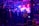 A group of people congregates under an industrial-style roof with metal beams, illuminated by vibrant purple and blue lighting. Smoke or mist partially obscures the area, adding to the atmospheric setting. A person in a bright yellow security jacket stands toward the foreground, and metal barricades are present along with large speakers.