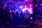 A group of people congregates under an industrial-style roof with metal beams, illuminated by vibrant purple and blue lighting. Smoke or mist partially obscures the area, adding to the atmospheric setting. A person in a bright yellow security jacket stands toward the foreground, and metal barricades are present along with large speakers.
