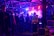 A group of people congregates under an industrial-style roof with metal beams, illuminated by vibrant purple and blue lighting. Smoke or mist partially obscures the area, adding to the atmospheric setting. A person in a bright yellow security jacket stands toward the foreground, and metal barricades are present along with large speakers.