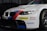 A white BMW race car prominently displaying sponsorship stickers including Dunlop, Castrol Edge, and Rays. The vehicle features sporty red and blue stripes running along the body, with a large front grille and sleek headlights adding to its aerodynamic and aggressive design.