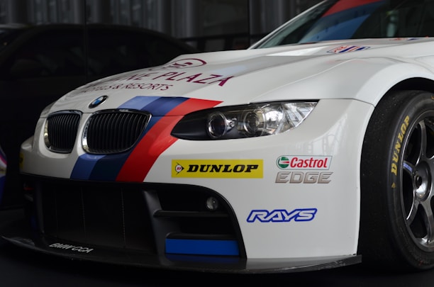 A white BMW race car prominently displaying sponsorship stickers including Dunlop, Castrol Edge, and Rays. The vehicle features sporty red and blue stripes running along the body, with a large front grille and sleek headlights adding to its aerodynamic and aggressive design.