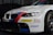 A white BMW race car prominently displaying sponsorship stickers including Dunlop, Castrol Edge, and Rays. The vehicle features sporty red and blue stripes running along the body, with a large front grille and sleek headlights adding to its aerodynamic and aggressive design.