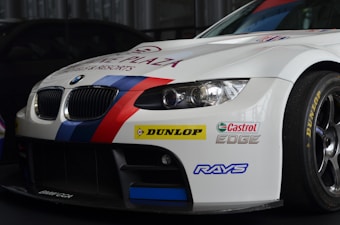 A white BMW race car prominently displaying sponsorship stickers including Dunlop, Castrol Edge, and Rays. The vehicle features sporty red and blue stripes running along the body, with a large front grille and sleek headlights adding to its aerodynamic and aggressive design.