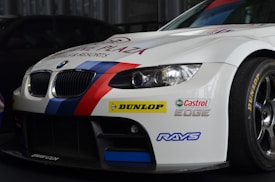 A white BMW race car prominently displaying sponsorship stickers including Dunlop, Castrol Edge, and Rays. The vehicle features sporty red and blue stripes running along the body, with a large front grille and sleek headlights adding to its aerodynamic and aggressive design.