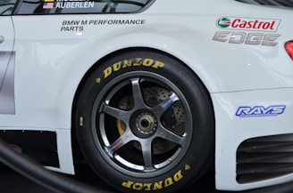 A close-up view of a sports car's rear wheel and partial body. The car has prominent sponsorship logos, including Castrol Edge and Dunlop, on a clean white surface. The wheel features a dark metallic rim and is equipped with Dunlop tires.