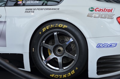 A close-up view of a sports car's rear wheel and partial body. The car has prominent sponsorship logos, including Castrol Edge and Dunlop, on a clean white surface. The wheel features a dark metallic rim and is equipped with Dunlop tires.