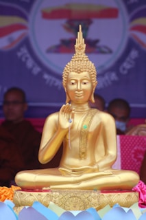 A golden statue of a seated figure with intricate detailing is prominently displayed. The figure is positioned in a meditative pose, with one hand raised. The background features blurred colors and possible text or design, suggesting a decorated setting.