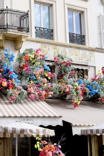 A charming facade of a building with ornate black wrought iron balconies and large windows. Below, vibrant, colorful flowers in shades of pink, red, blue, and yellow are arranged along a striped awning. The awning displays the name 'Le Bonheur' and features beige and white stripes. The building exudes a lively, inviting atmosphere with its floral decorations and classic architectural elements.
