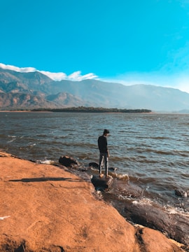 A person stands at the edge of a rocky shoreline, gazing out at a large body of water. The sky is clear and blue, with mountains visible in the distance. The landscape conveys a sense of vastness and tranquility.