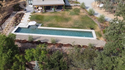 An aerial view of a backyard featuring a rectangular swimming pool with a stone walkway leading to a shaded area with an umbrella. The surrounding landscape includes grass areas, shrubs, and a patio adjacent to a modern house. The scene is bordered by trees and a gravel pathway.