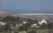 A serene landscape featuring a hillside covered in lush green trees and sparse vegetation. In the foreground, a small village with white-washed buildings and a church with a distinctive blue dome stands out amidst the greenery. Beyond the village, a vast expanse of flat farmland stretches towards a calm blue sea, with islands visible in the distance under a clear sky.