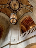 An ornate architectural interior features an intricately decorated ceiling with gold and patterns. A vintage chandelier hangs prominently, casting a warm glow over the marble archways and detailed moldings. The overall design reflects a classic and grand style.