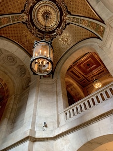 An ornate architectural interior features an intricately decorated ceiling with gold and patterns. A vintage chandelier hangs prominently, casting a warm glow over the marble archways and detailed moldings. The overall design reflects a classic and grand style.