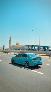 A turquoise-colored car is driving on a multi-lane highway under a clear blue sky. In the background, a gas station is visible on the right side of the road, along with a mosque that features a dome and a tall minaret.