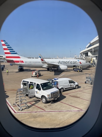 A large airplane is parked at the airport terminal. It has the branding and tail colors of an airline. Several service vehicles and staff are present on the tarmac, including vans and maintenance equipment. The scene is viewed through an airplane window with part of the airport structure visible.