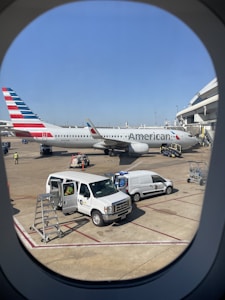 A large airplane is parked at the airport terminal. It has the branding and tail colors of an airline. Several service vehicles and staff are present on the tarmac, including vans and maintenance equipment. The scene is viewed through an airplane window with part of the airport structure visible.