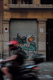 A street art piece on a metal shutter featuring a cartoonish green dragon with orange eyes and a crown, alongside a lit cigarette. A person wearing a red helmet is riding a motorcycle, creating a motion blur across the image.