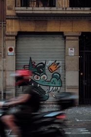 A street art piece on a metal shutter featuring a cartoonish green dragon with orange eyes and a crown, alongside a lit cigarette. A person wearing a red helmet is riding a motorcycle, creating a motion blur across the image.