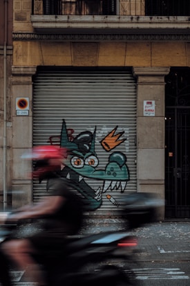 A street art piece on a metal shutter featuring a cartoonish green dragon with orange eyes and a crown, alongside a lit cigarette. A person wearing a red helmet is riding a motorcycle, creating a motion blur across the image.