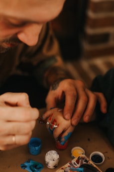 A close-up of an adult hand guiding a child's hand as they apply paint with a small brush. The child's hand is already covered in blue and red paint, and several small paint pots in various colors are visible nearby. The setting appears to be a warm, cozy environment.