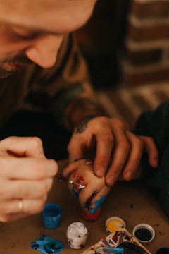 A close-up of an adult hand guiding a child's hand as they apply paint with a small brush. The child's hand is already covered in blue and red paint, and several small paint pots in various colors are visible nearby. The setting appears to be a warm, cozy environment.