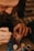 A close-up of an adult hand guiding a child's hand as they apply paint with a small brush. The child's hand is already covered in blue and red paint, and several small paint pots in various colors are visible nearby. The setting appears to be a warm, cozy environment.