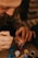 A close-up of an adult hand guiding a child's hand as they apply paint with a small brush. The child's hand is already covered in blue and red paint, and several small paint pots in various colors are visible nearby. The setting appears to be a warm, cozy environment.