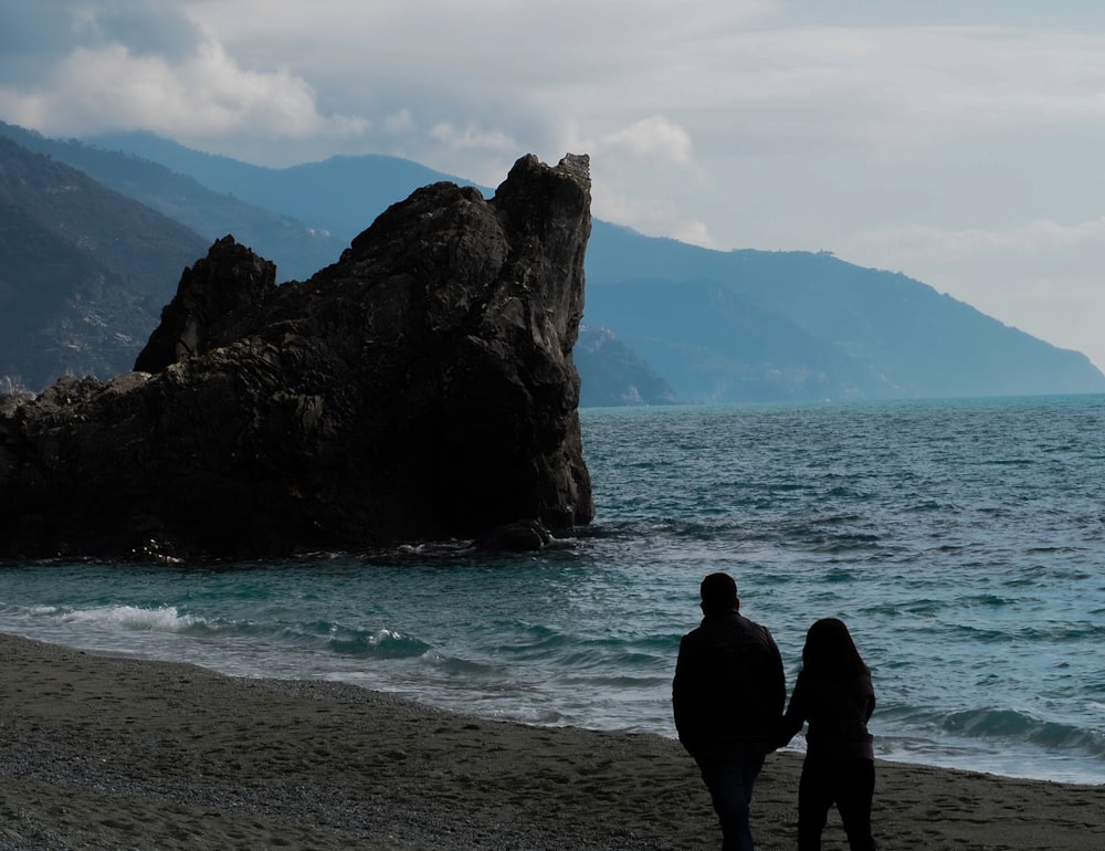 a couple people standing on a beach looking at a large rock formation